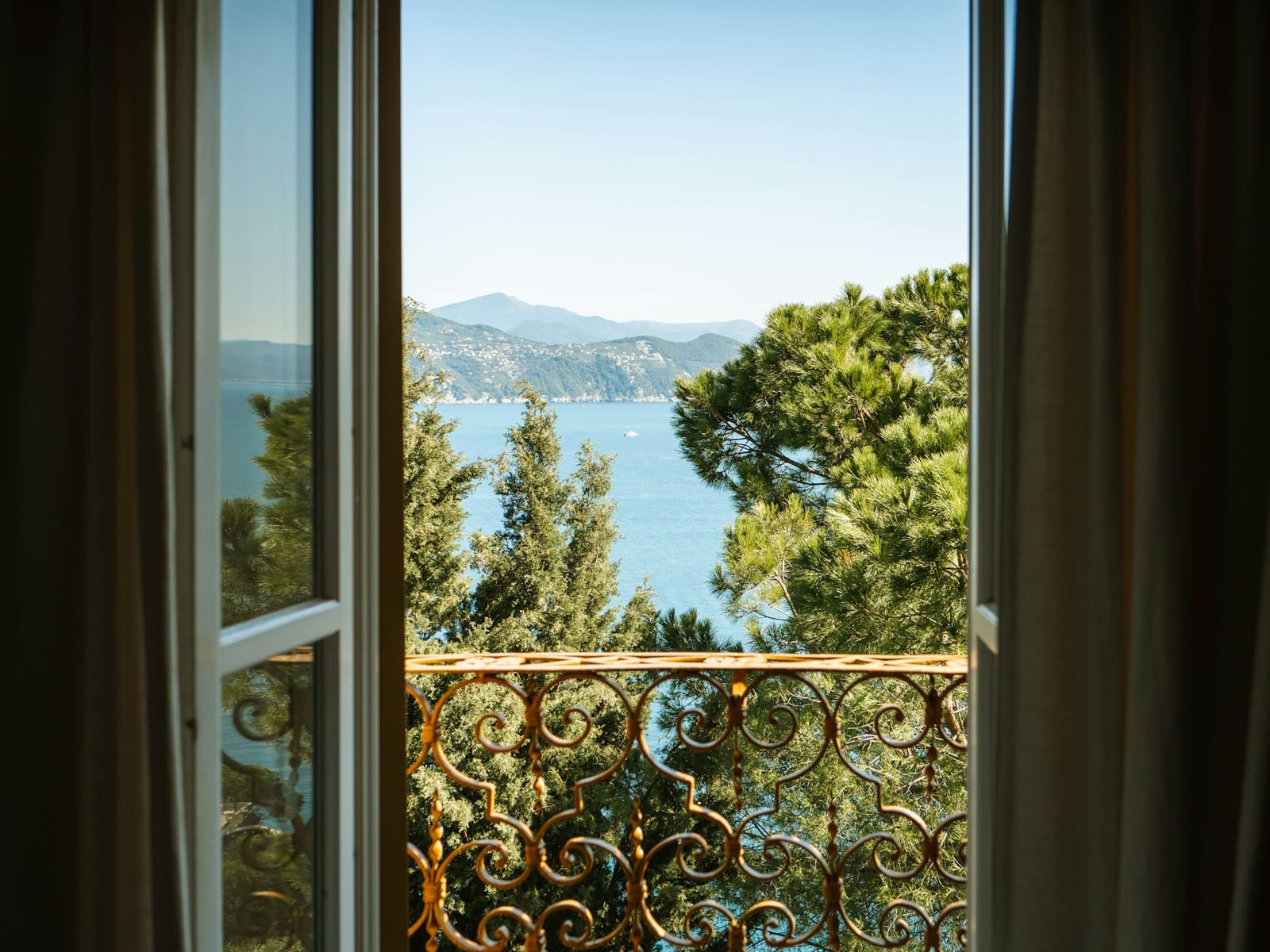 An open window with a balcony rail and a view of trees and sea